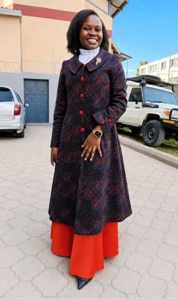 A woman standing outdoors in a long patterned coat-dress buttoned to a high collar, worn over a full-length red skirt, with a white high-necked blouse beneath.