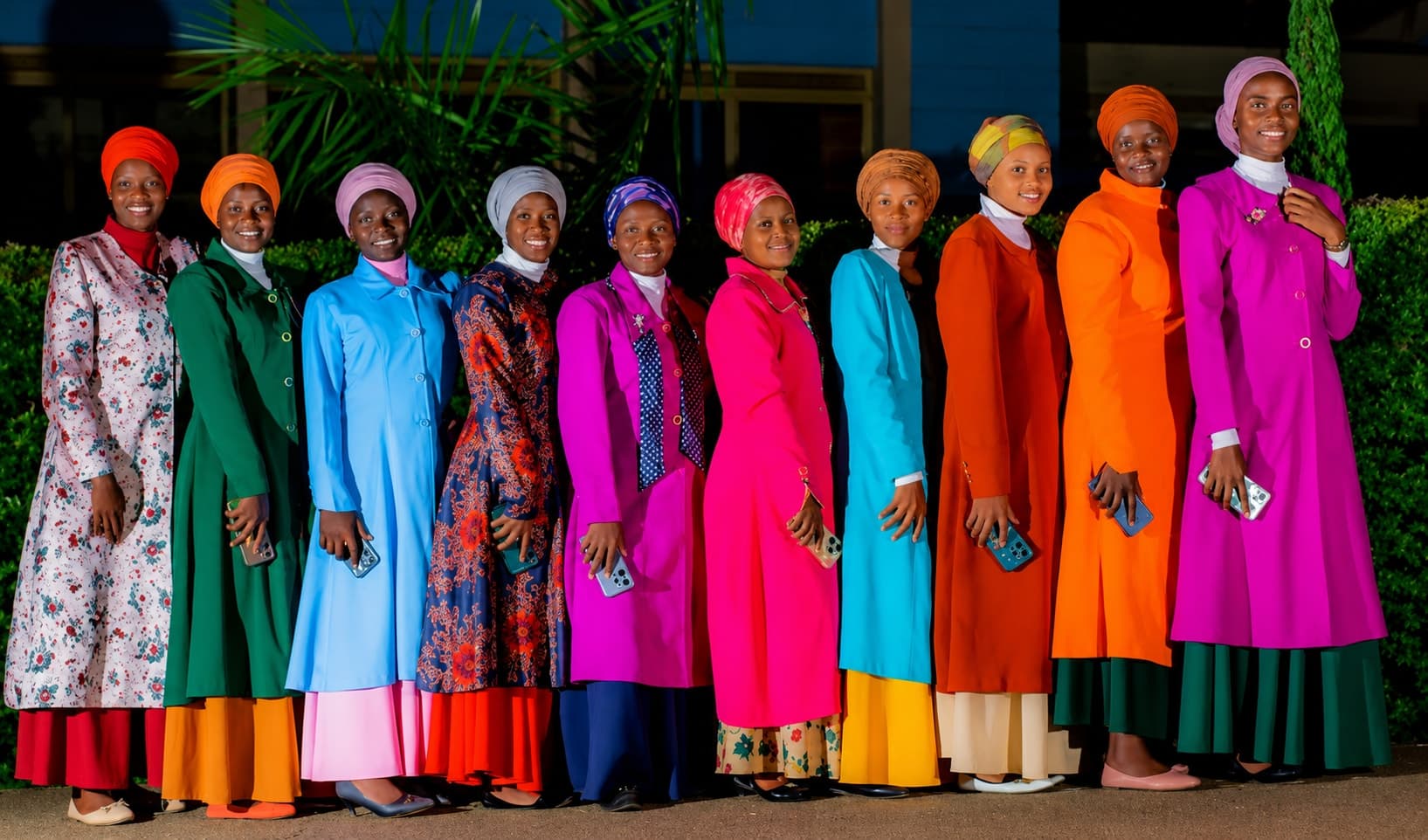 Ten women standing in a line in ankle-length coats and dresses — floral, green, blue, turquoise, orange, red, magenta — each with a headscarf and a high collar, smiling towards the camera.