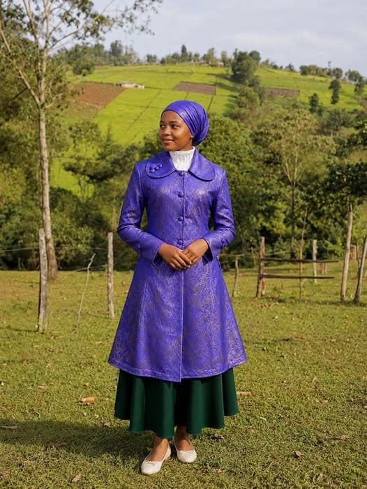 A woman standing on grass in a long purple brocade coat over an ankle-length dark green skirt, with a matching purple headscarf and a high white collar, tea-covered hills behind her.