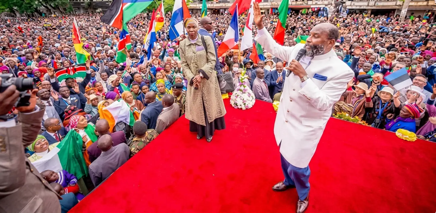 Prophet David Owuor in a white robe on a red carpet, one hand raised, addressing a very large crowd holding the flags of many nations.