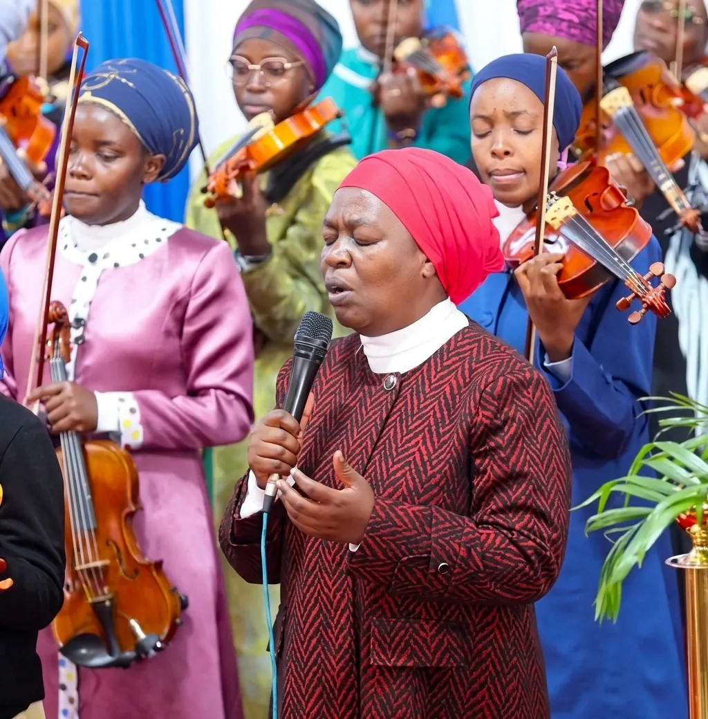 A woman in a red head covering singing into a microphone, violinists standing behind her.