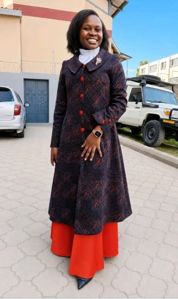 A woman standing outdoors in a long patterned coat-dress buttoned to a high collar, worn over a full-length red skirt, with a white high-necked blouse beneath.