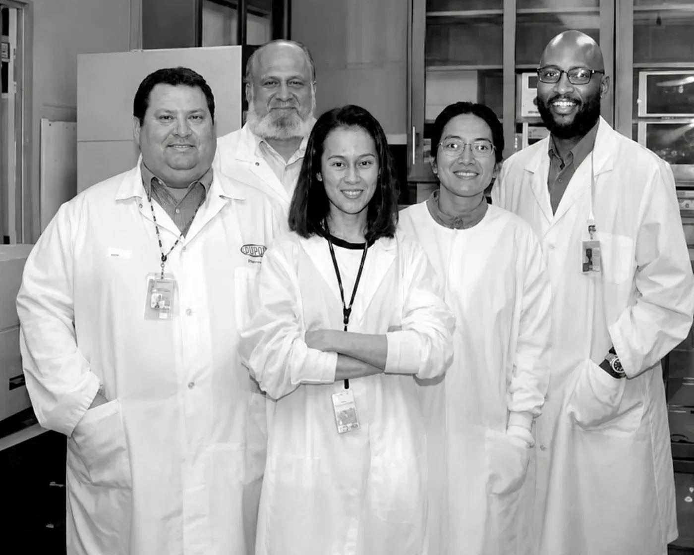Five colleagues in white laboratory coats standing together for a group photograph in a research laboratory, cabinets and equipment behind them.