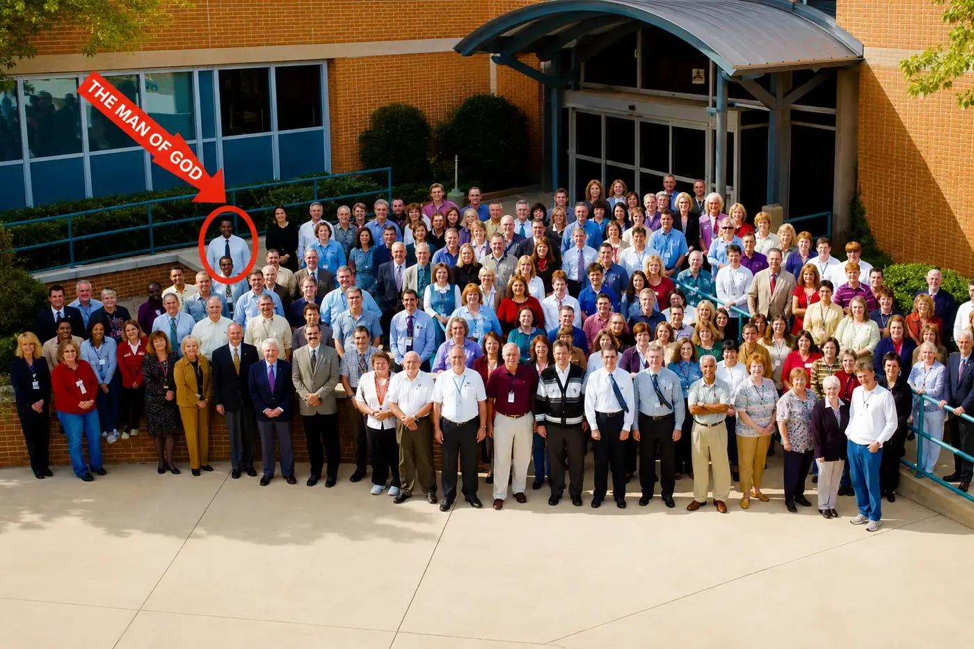 A large institutional staff photograph of several hundred people arranged on steps outside a brick building, with a red arrow and circle marking one man near the back left.