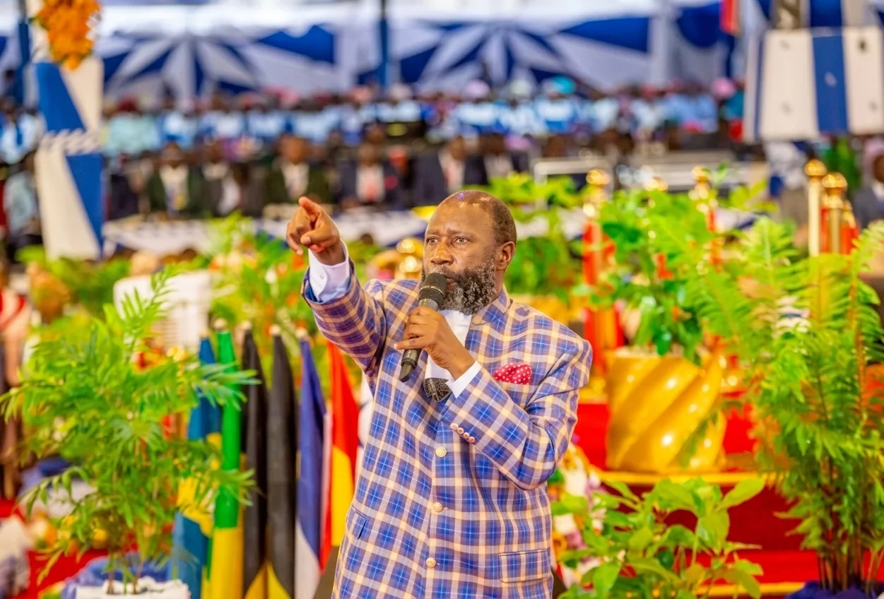 Prophet David Owuor preaching with a microphone, one arm outstretched, before tiered rows of seated clergy, with national flags and greenery along the front of the platform.