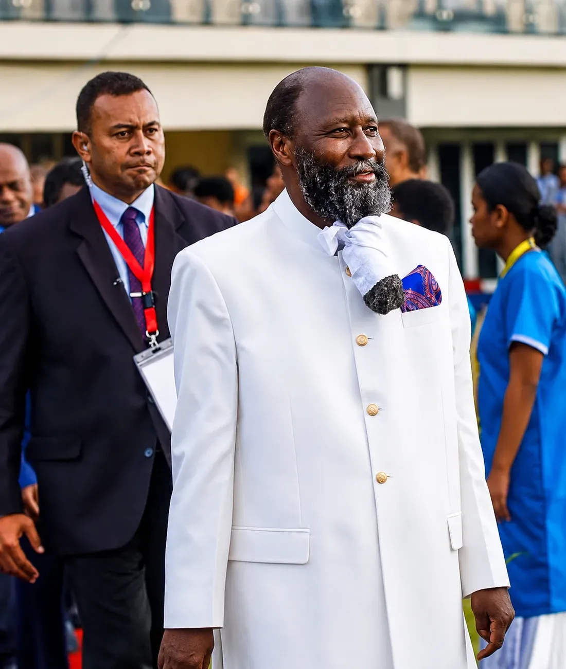 Prophet David Owuor in a white robe, walking through a crowd at an outdoor gathering, accompanied by an official carrying a clipboard.