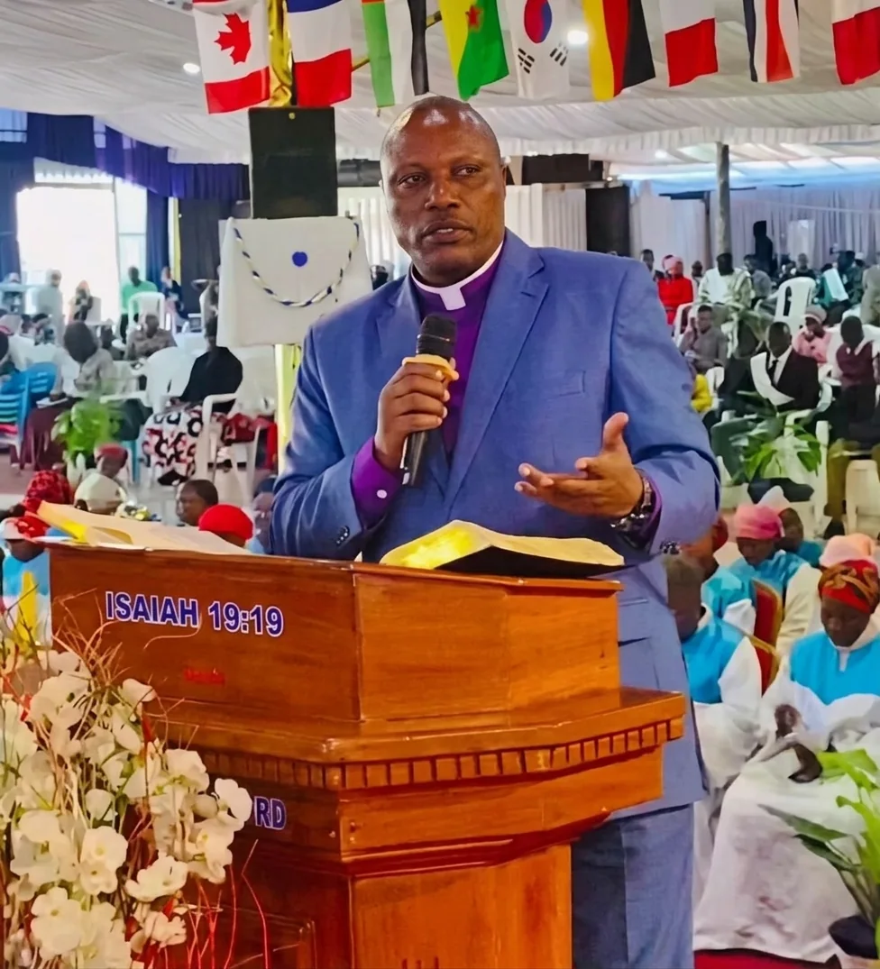 A preacher in a blue suit speaking from a wooden pulpit marked ISAIAH 19:19, an open Bible before him and the flags of many nations hanging above.