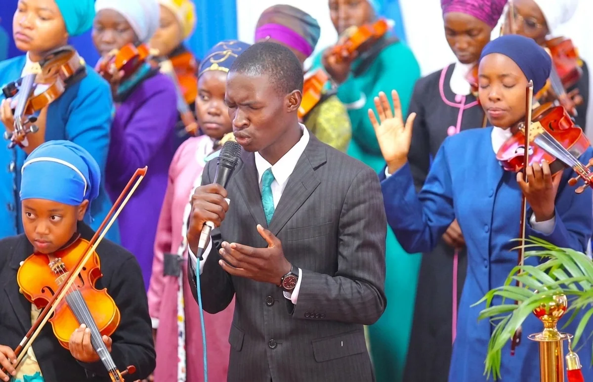 A young man singing into a microphone at the front of a congregation, with a string orchestra of women in headscarves playing violins behind him.
